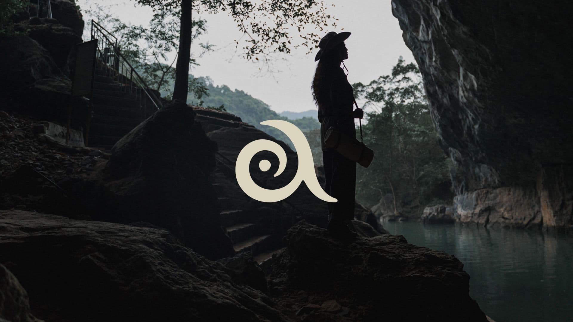 Silhouette of a traveler overlooking a tranquil river inside a rocky cave, overlaid with the Auko logo.